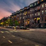 City street lined with historic brownstone buildings at sunset, glowing windows, parked cars, and trees along the sidewalk.