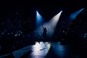 Singer standing on a stage under bright lights with a crowd in the background.