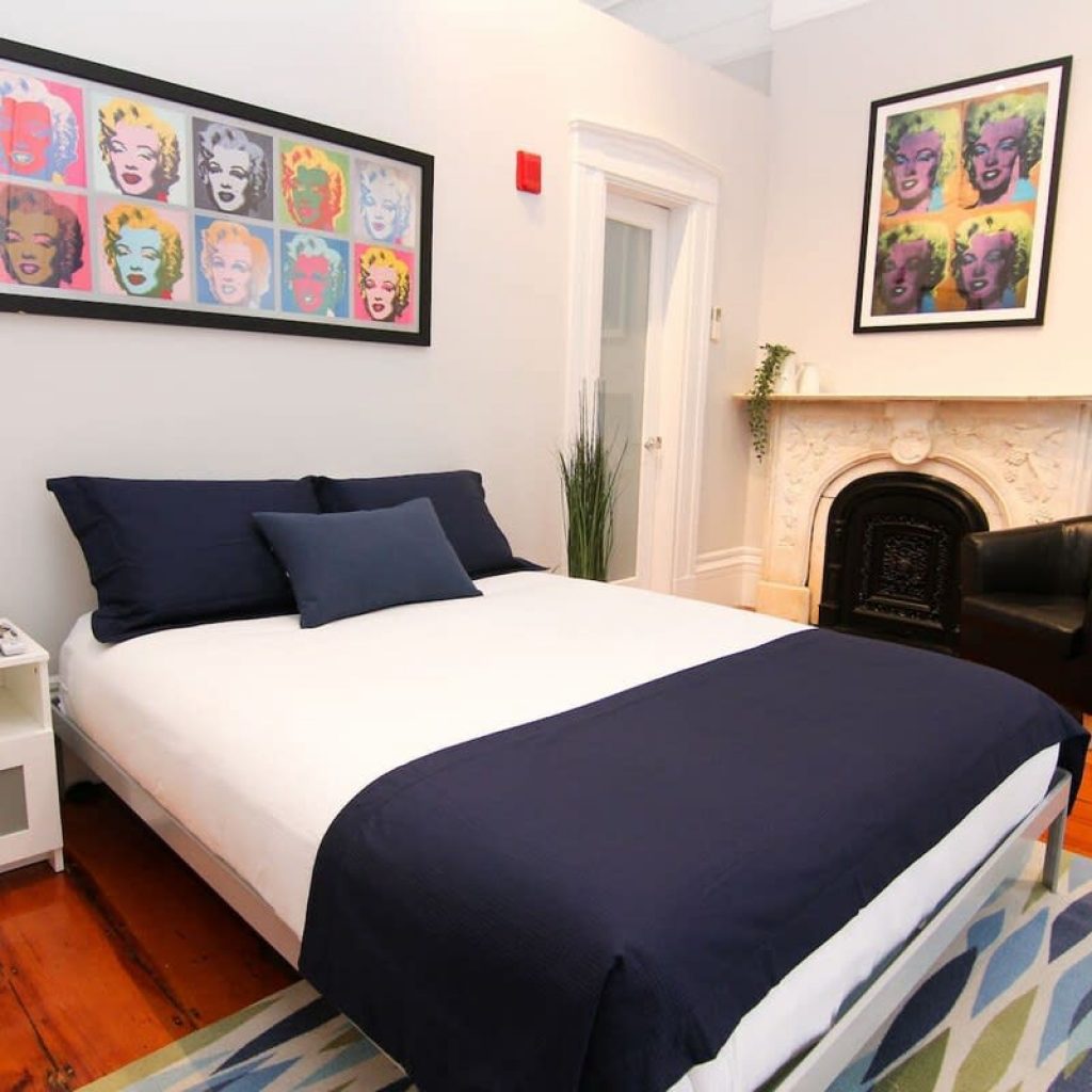 Modern bedroom with a white bed, navy bedding, pop art prints, fireplace, armchair, and wooden floors.