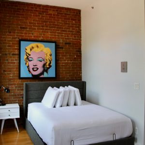 Modern bedroom with white bed, brick accent wall, pop art portrait, bedside table, and wooden floor