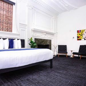 Spacious modern bedroom with a large bed, high ceilings, fireplace, two black chairs, wall art, and a mounted TV.