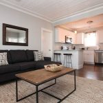Cozy living room with a dark sofa, wooden coffee table, rug, and an open kitchen with white cabinets and stools.